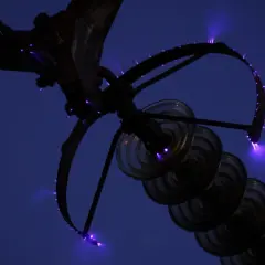 Des arcs électriques violets, les feux de Saint-Elme, crépitent sur une structure métallique et des isolateurs en verre contre un ciel nocturne.