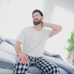 Uomo con torcicollo dopo aver dormito, si massaggia il collo con espressione sofferente, seduto sul letto.