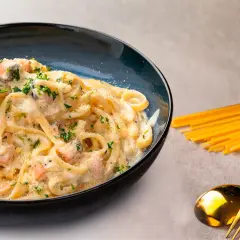 Delicioso plato de pasta con salmón y gambas, adornado con perejil fresco y queso rallado. Acompañado de espaguetis crudos y cubiertos dorados.