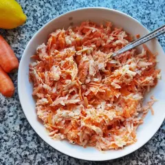 Pyszna surówka z marchewki jak z restauracji, gotowa do podania. Obok leżą świeże marchewki i cytryna.