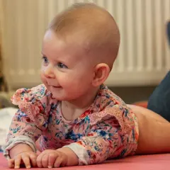 Uśmiechnięta dziewczynka w kwiecistym body leży na macie, wykonując próbę trakcji. Obok siedzą dorośli.