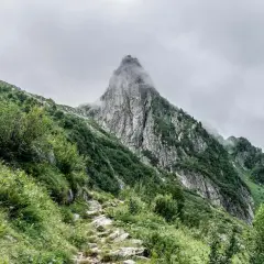Sentier rocailleux grimpant vers un pic montagneux enveloppé de brume. Végétation luxuriante sur les pentes.