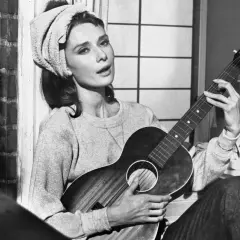 Audrey Hepburn, z ręcznikiem na głowie, gra na gitarze, śpiewając "Moon River".