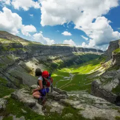 Senderista contempla el valle verde y las imponentes paredes rocosas de Monte Perdido, Huesca, bajo un cielo azul con nubes.