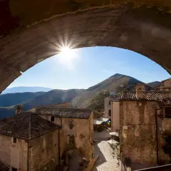 Solstrålar bryter fram genom en stenbåge över en pittoresk by i Abruzzo, Italien. Kulliga berg i bakgrunden.