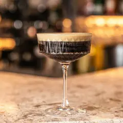 Un bicchiere di caffè martini con schiuma cremosa su un bancone di marmo, con luci sfocate sullo sfondo.