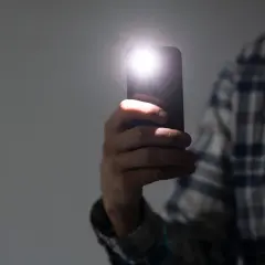 Dłoń trzyma telefon, którego lampa błyskowa rozświetla ciemność. Dowiedz się, jak włączyć lampę błyskową w telefonie, by zawsze mieć światło.