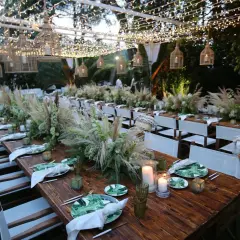 Tavoli in legno apparecchiati per un matrimonio in campagna, con centrotavola di fiori e pampas, luci soffuse e cesti decorativi.