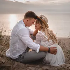 Romantyczna sesja we dwoje na plaży. Para siedzi blisko siebie, otoczona trawami, z widokiem na spokojne morze o zachodzie słońca.