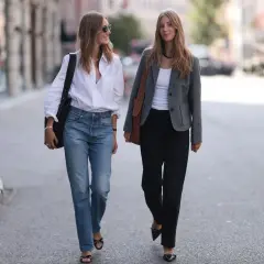Dos mujeres caminan por la calle. Una, con un estilo demure, lleva vaqueros y camisa blanca. La otra, con chaqueta gris y pantalones negros, camina con elegancia.
