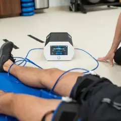 Pacjent leży na macie, podłączony do urządzenia emitującego prądy na kolano. Terapeuta czuwa nad przebiegiem rehabilitacji.