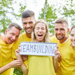 En grupp glada människor i gula tröjor håller upp en skylt med "TEAMBUILDING". De verkar ha det roligt, som om de precis svarat på några snabba roliga frågor.