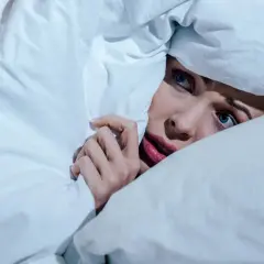 Verängstigte Frau versteckt sich unter der Decke, die Augen weit aufgerissen, als ob sie einen Albtraum erlebt.