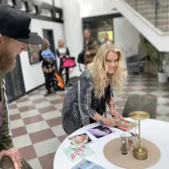 Ankie Bagger signerar bilder vid ett bord. En man med skägg och keps står bredvid.