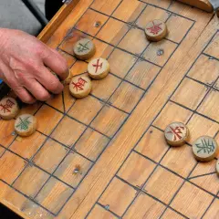 Eine Hand bewegt einen Stein beim China Schach. Das Holzbrett ist aufgeschlagen, die Figuren mit chinesischen Schriftzeichen sind verteilt.