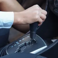 Dłoń na dźwigni **automatycznej skrzyni biegów**. Widać oznaczenia P, R, N, D, 3, 2, L.