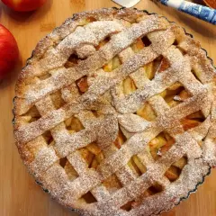 Pyszna tarta z jabłkami, posypana cukrem pudrem, leży na drewnianej desce obok świeżych jabłek.