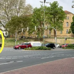 Eine Person wartet an der Ampel, um links abzubiegen. Autos stehen bereit, während im Hintergrund ein Gebäude und Bäume zu sehen sind.