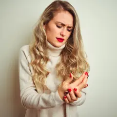 Kobieta z blond włosami trzymająca się za bolącą rękę, ubrana w beżowy golf.