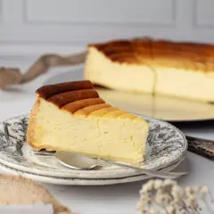 Une part de tarte au fromage, dorée et crémeuse, sur une assiette ornée. Le reste du gâteau est visible en arrière-plan.