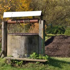 Stary, betonowy krąg studni kręgowej z metalową korbą i liną, otoczony jesienną zielenią.