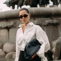 Elegancka kobieta w białej koszuli i okularach przeciwsłonecznych, z czarną torebką. Stylizacja godna polskiego domu mody.