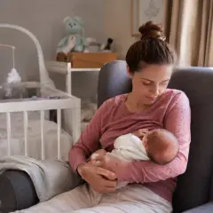 Matka karmi piersią noworodka w fotelu. W tle widać łóżeczko dziecięce i pluszowego misia. To piękny obrazek z okresu po porodzie.