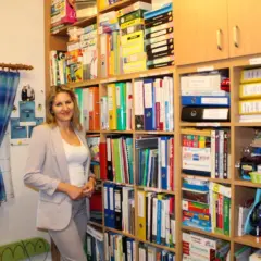 Kobieta w jasnej marynarce stoi w nowoczesnym gabinecie logopedycznym, otoczona regałami pełnymi materiałów terapeutycznych.