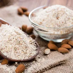 Cuillère en bois remplie de poudre d'amande, idéale pour remplacer la farine dans vos recettes. Amandes entières et bol de poudre à côté.