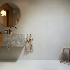 Modernt badrum med handfat i marmor och väggar i microcement. En rund spegel och två lampor ovanför.