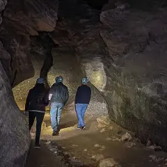 Tres personas con cascos exploran una cueva oscura, sus linternas iluminan las paredes rocosas y el suelo irregular. Los yesos naturales brillan en la oscuridad.