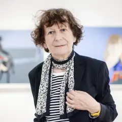 Ewa Kuryluk, artystka o burzy brązowych włosów, w czarnej marynarce i pasiastym szalu, pozuje na tle swoich obrazów.