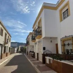 Calle soleada con casas blancas y detalles amarillos en Celin, Almería. Balcones de hierro forjado y un cielo azul con nubes.