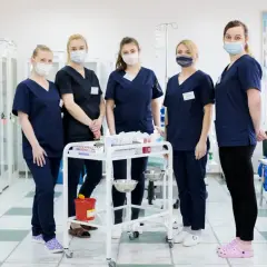 Grupa studentek pielęgniarstwa w maseczkach stoi przy wózku medycznym w sali szpitalnej.