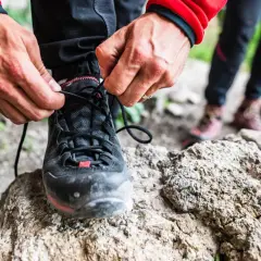 Dłonie zawiązują sznurówki czarnych butów podejściowych, gotowych na górską wyprawę. W tle widać inne buty trekkingowe.