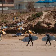 Deux surfeurs marchent sur la plage de Tamraght, Maroc, avec leurs planches sous le bras. Des parasols et des chaises longues sont installés sur le sable.