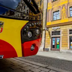 Żółto-czerwony autobus 143 Kraków z herbem miasta na błotniku, zaparkowany przed kamienicą z malowidłami i sklepem Żabka.