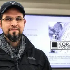 Man med skägg och keps framför en skärm med texten "KORAN PODDEN". Han ser tacksam ut, som om han ber: "ربي اوزعني ان اشكر نعمتك".