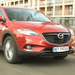 Czerwona Mazda CX-9 w ruchu miejskim, z widocznym kierowcą za kierownicą.