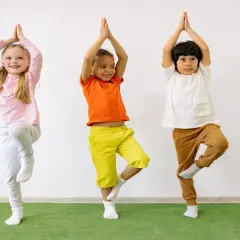 Tre bambini praticano yoga, in piedi su una gamba sola, con le mani giunte sopra la testa.