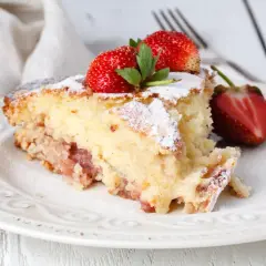 Puszyste ciasto na maślance z truskawkami, posypane cukrem pudrem, na białym talerzu.