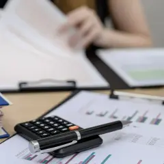 Na biurku leży kalkulator i długopis na wykresach. W tle osoba przegląda dokumenty, być może analizując podatek od najmu.
