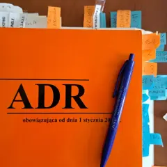 Pomarańczowa okładka z napisem ADR i niebieski długopis. Dokumenty z zakładkami, w tym te dotyczące uprawnień ADR.
