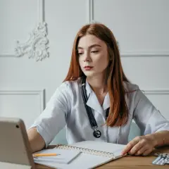 Młoda lekarka z rudymi włosami, w białym fartuchu, z notatnikiem i tabletami, szuka informacji o hemoroidach jaki lekarz.