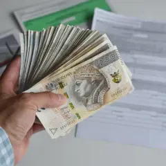 Dłoń trzyma plik banknotów 200 zł, symbolizujących dochód netto, na tle dokumentów podatkowych.