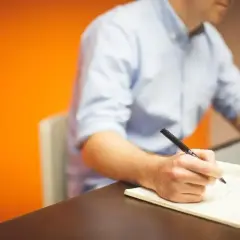 Mężczyzna w niebieskiej koszuli pisze notatki w zeszycie, pracując przy laptopie. Daje mu to **prokura** do działania.