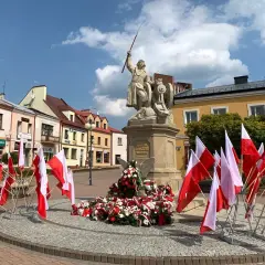 Pomnik z polskimi flagami, w tym kierunkowy 15, na placu przed kolorowymi kamienicami.