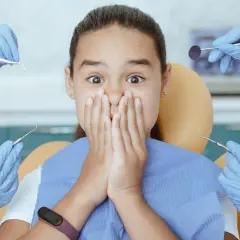 Przerażona dziewczynka w gabinecie dentystycznym, otoczona rękami w rękawiczkach z narzędziami. Czy to już moment, kiedy wychodzą 5 zębów u dziecka?