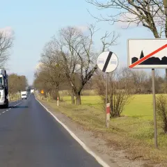 Znaki drogowe informują, ile można jechać poza terenem zabudowanym. Ciężarówka jedzie drogą, mijając pola i drzewa.
