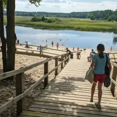 Dziewczynka schodzi drewnianymi schodami na piaszczystą plażę nad jeziorem. W oddali widać ludzi kąpiących się i odpoczywających. Stawy echo zwierzyniec to idealne miejsce na letni relaks.